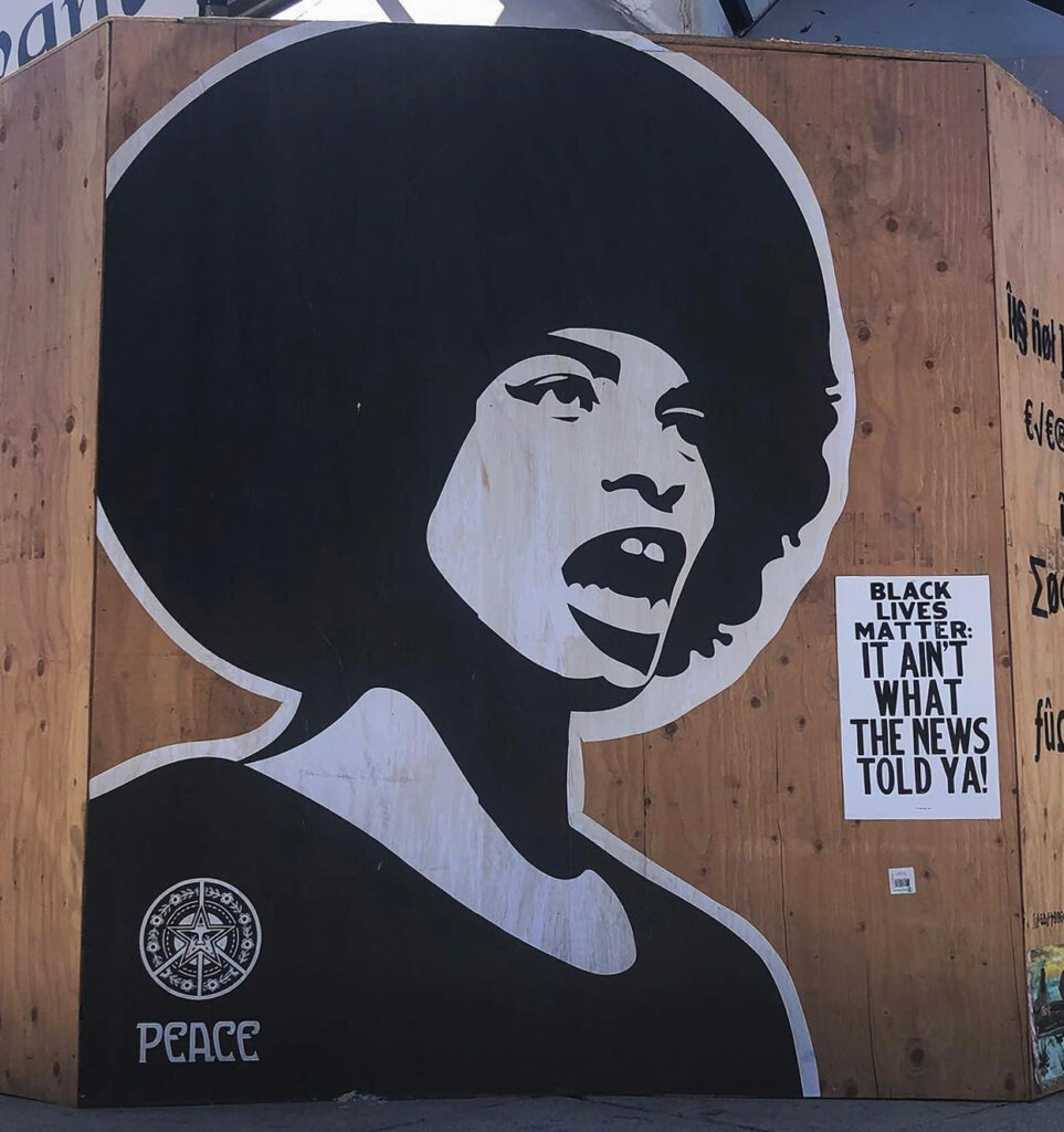 A brown plywood wall with a black and white wheat-paste poster of a figure mid-sentence placed next to a rectangular poster with text that reads, "Black Lives Matter: It Ain't What The News Told Ya!" in all-caps bold, black type.